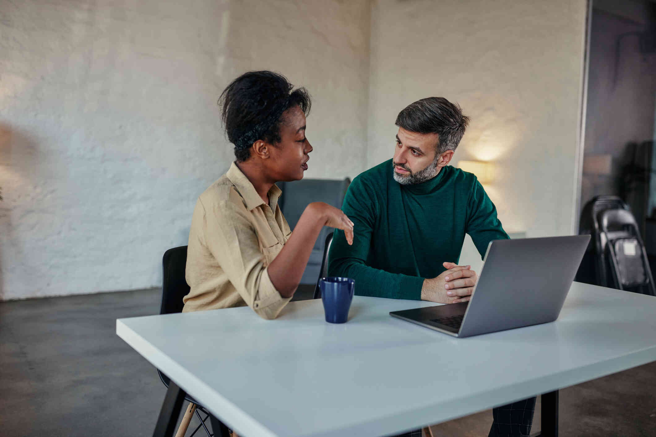 A man and woman sits next to each other at a table that hasna laptop and coffee mug on it as they talk to each other with serious expressions.